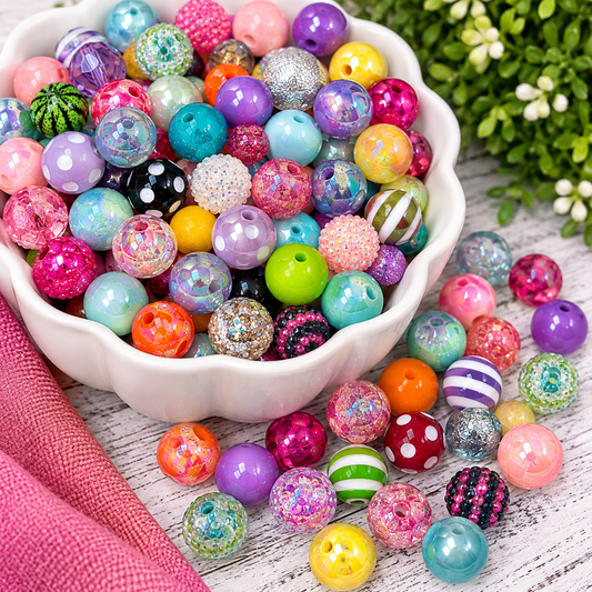 Colorful assortment of acrylic craft beads featuring pearl, glitter, rhinestone, striped, polka dot and iridescent finishes displayed in a glass bowl with scattered beads.