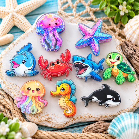 Colorful ocean animal acrylic focal beads featuring dolphins, jellyfish, starfish, crabs, sharks, sea turtles, seahorses, and orcas displayed on a coastal-themed wooden tray. Sold in randomly assorted bags of 6.