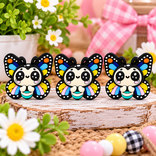 Cute rainbow butterfly silicone focal bead with black wings decorated in colorful blue, yellow, pink, and orange details, large sparkling eyes, rosy cheeks, and turquoise striped body, displayed on a rustic wooden tray with spring flowers.