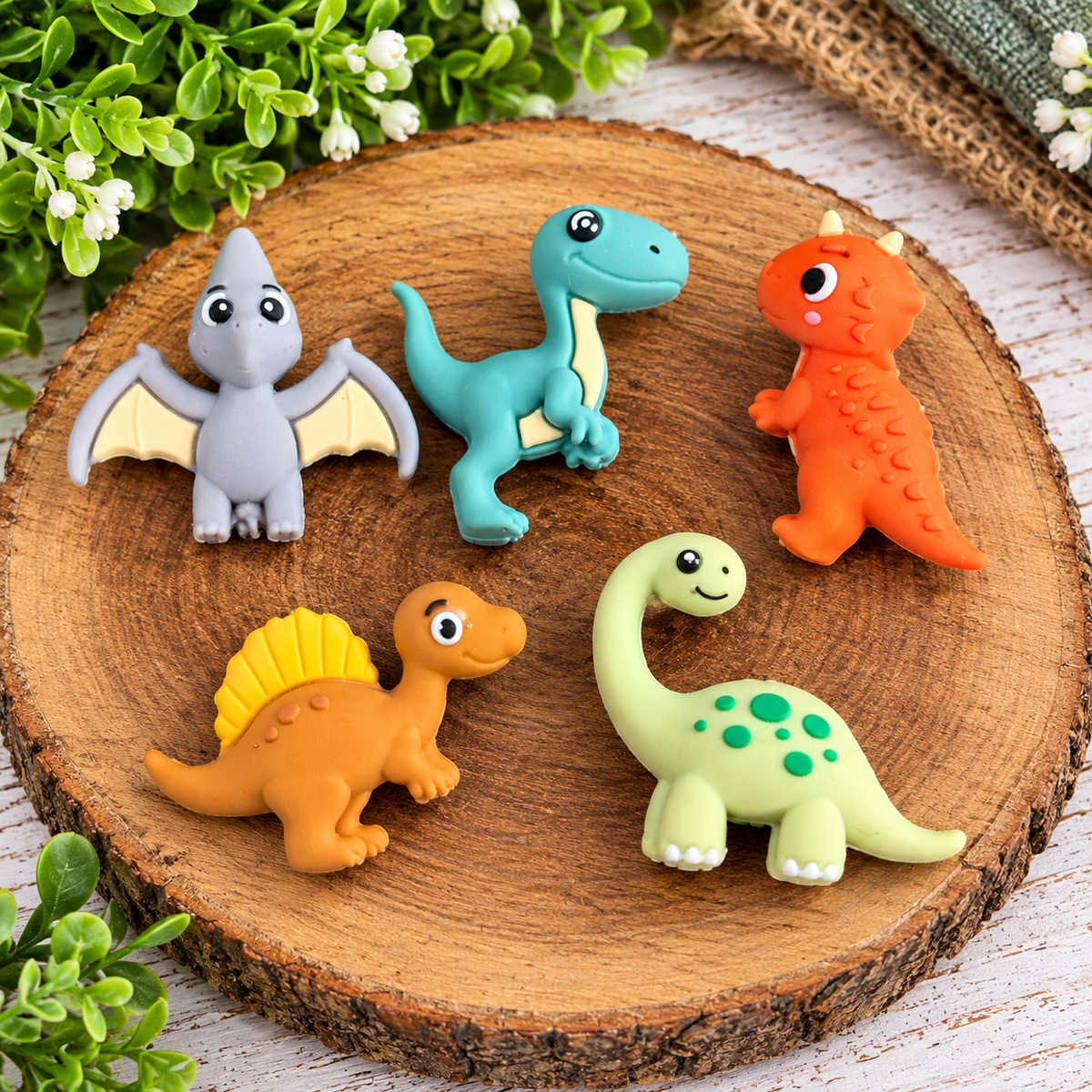 Five colorful dinosaur silicone focal beads featuring a gray flying pterodactyl, teal raptor, orange dinosaur, brown dinosaur with yellow back sail, and green spotted long-neck dinosaur displayed on a rustic wooden tray.